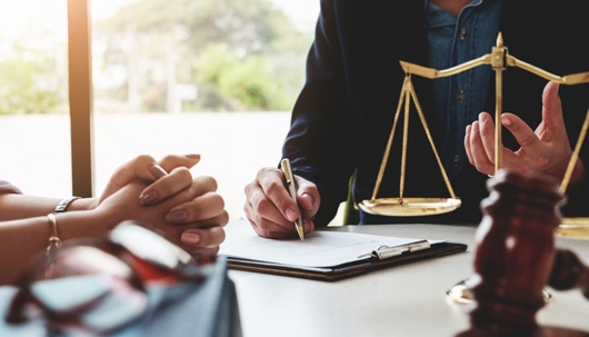 A lawyer consults with a client at a table with legal documents, a gavel, and scales of justice.