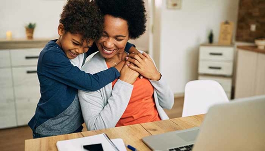 A woman smiles as a child hugs her from behind while she works on a laptop at a table.