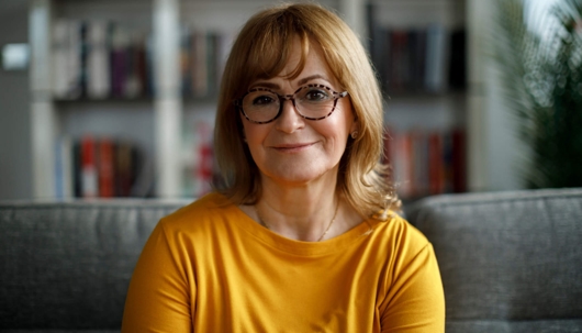 A smiling woman with glasses and a yellow shirt sits on a couch in front of a bookshelf.