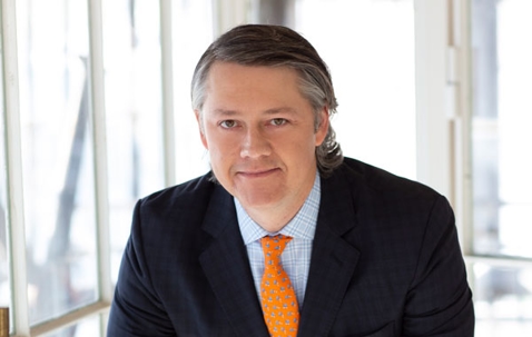 Professional headshot of a man with graying hair wearing a suit and bright orange tie.