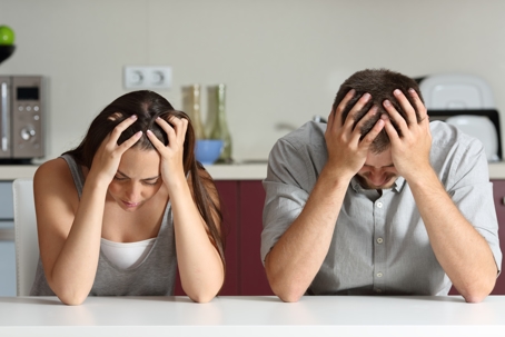 A couple sits dejectedly at a table, heads in their hands.