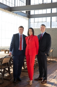 Three individuals pose indoors, one in a red suit and two in dark suits with ties.
