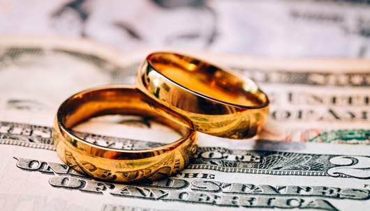 Two gold wedding bands rest on a stack of U.S. dollar bills.