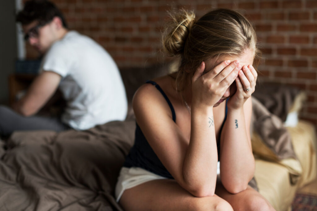A man and a woman in a bedroom sitting on opposite sides of the bed. The woman has her head in her hands while the man looks over his shoulder in her direction. The image visually conveys a couple fighting and on the verge of divorce or separation.